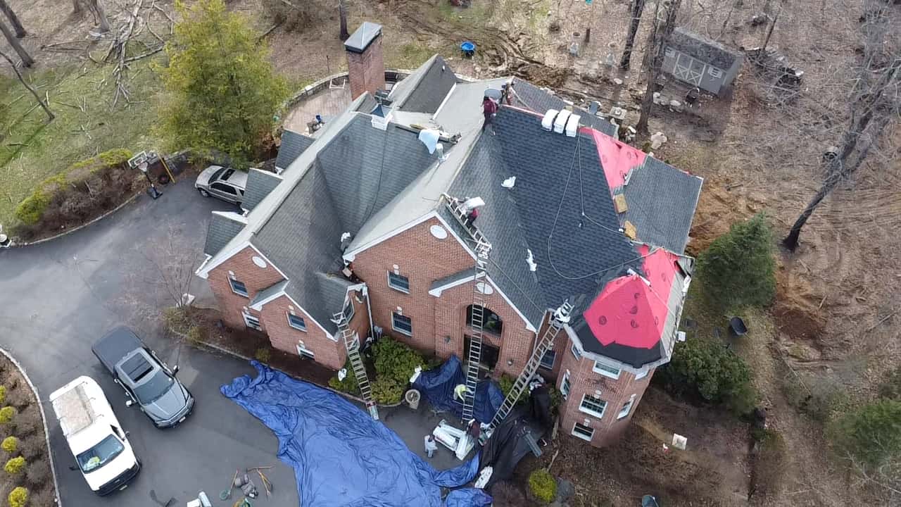 Aerial of a large brick and stucco home mid re-roof, red underlayment over the turret, new gray shingles on other slopes, crew and ladders present